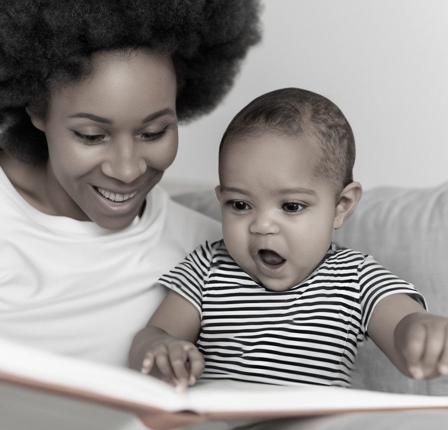 Eine glückliche Mutter mit ihrem Baby auf dem Schoß, die ihm ein Buch vorliest. Das Baby zeigt großes Interesse und zeigt mit dem Finger auf ein Bild im Buch